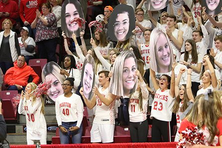 Morton High School fans hold enlarged player head signs in the crowd while cheering.