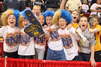 Notre Dame fan wear blue and gold wigs while waving foam finger in crowd.